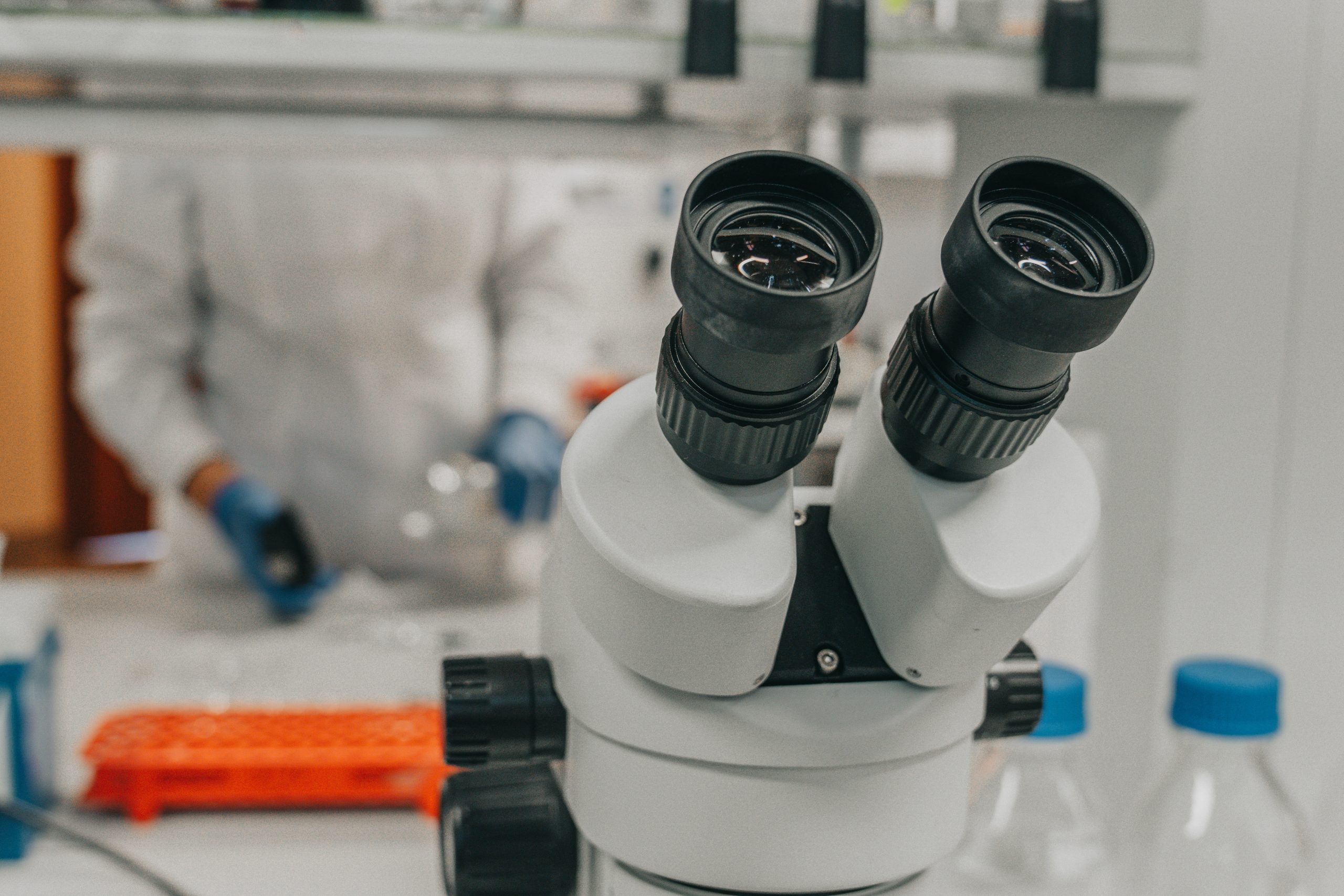 Laboratorium badawcze. Okular mikroskopu na stole laboratoryjnym. Osoba w fartuchu stojąca w tle.
