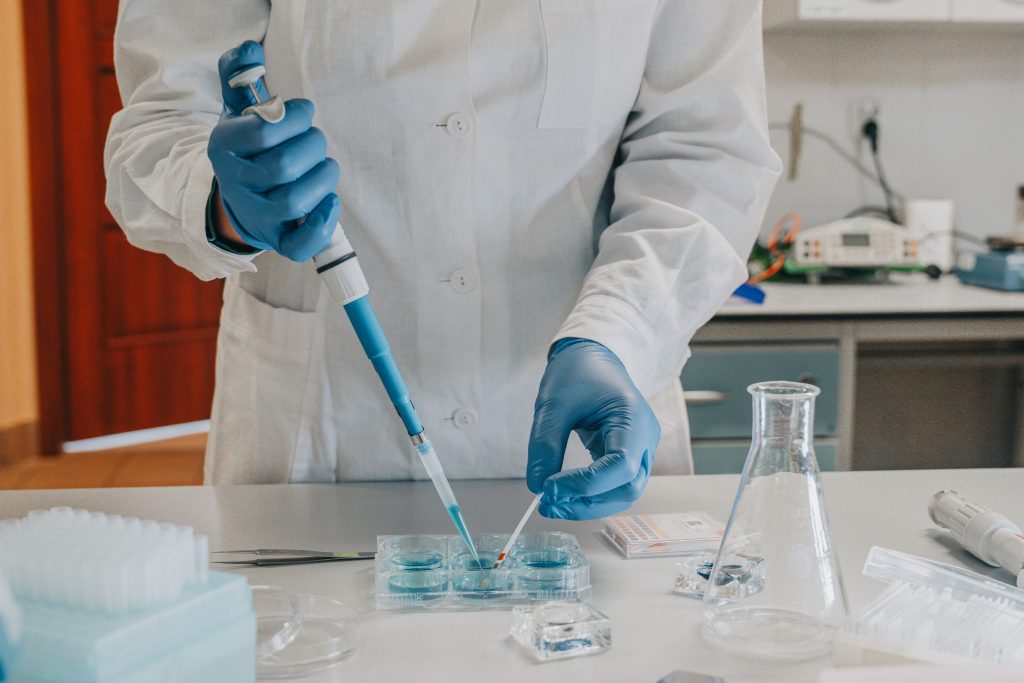 Osoba w fartuchu laboratoryjnym i rękawiczkach. Pipetuje. Widoczne naczynia laboratoryjne